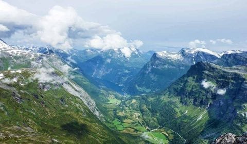 Panoramautsikt fra Dalsnibba over den grønne dalen, Geiranger og Geirangerfjorden, snø på fjellene og skyer som nærmer seg