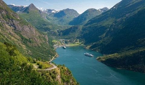 Hohe grüne Berge rund um den Geirangerfjord, wo zwei Schiffe im Hafen liegen, Blick von der Adlerstraße