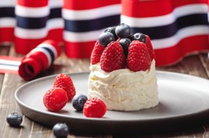 A small pavlova cake adorned with red and blue berries in front of a Norwegian flag to celebrate Syttende Mai.
