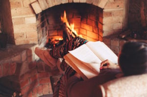 A woman reading a book by a fireplace inside of a cabin