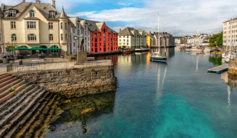View of Brosundet Ålesund, Norway, featuring colorful buildings along the water, boats docked in the harbor, and a statue in the foreground.